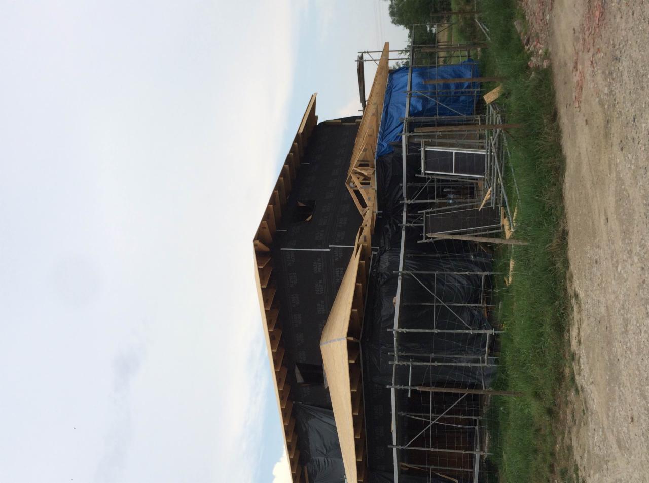 Budowa domu: konstrukcja szkieletowa z widocznym deskowaniem dachu i rusztowaniem, zabezpieczona czarną folią i plandeką.