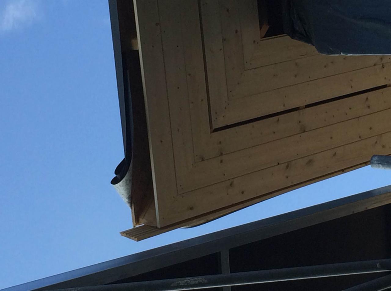 Fragment konstrukcji drewnianej, prawdopodobnie zadaszenia, widziany od dołu na tle błękitnego nieba, z widocznymi elementami metalowej ramy.