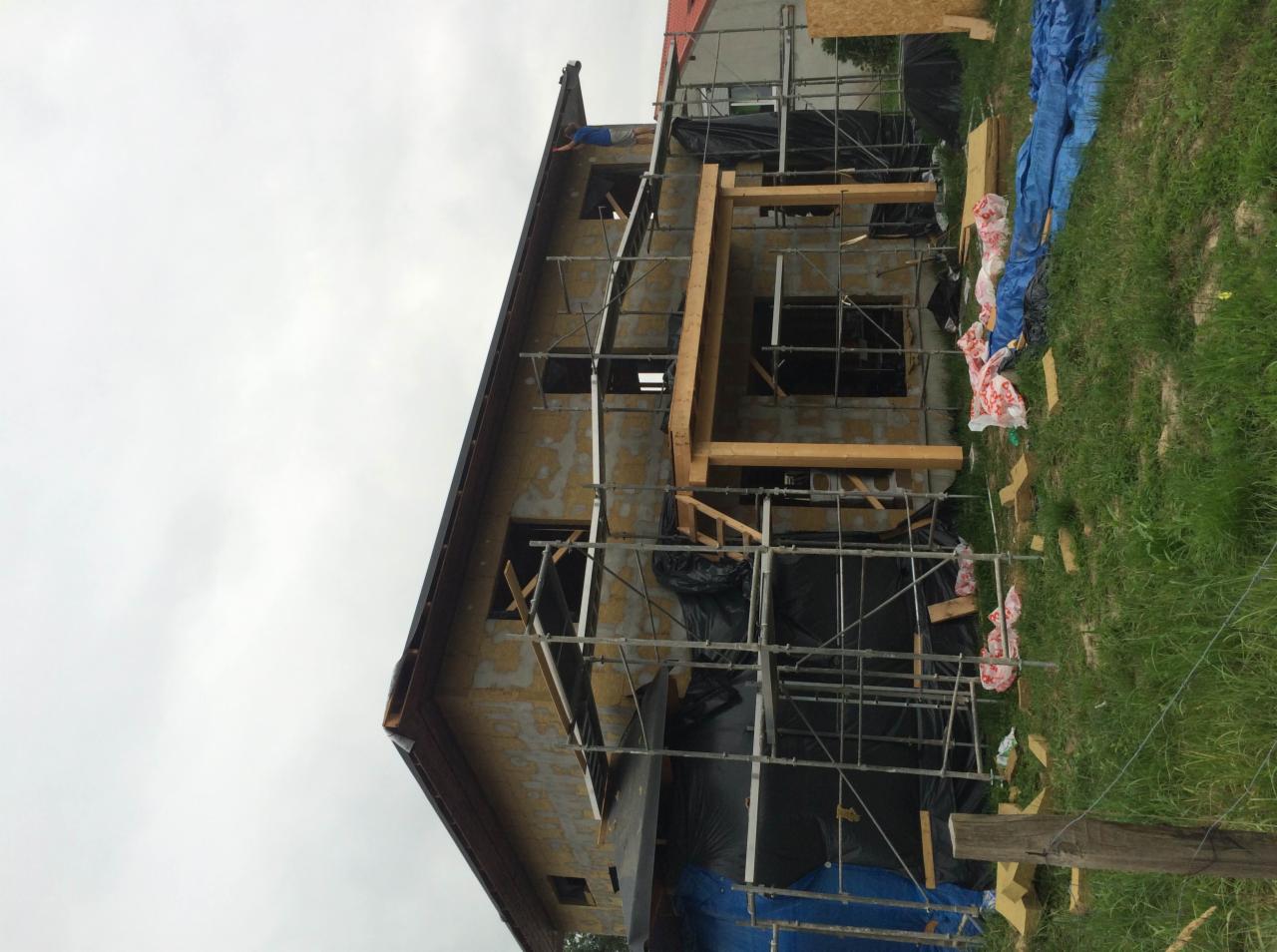 Budowa domu w trakcie wznoszenia, widoczne rusztowania, drewniane elementy konstrukcyjne i folia ochronna, pracownik na dachu.