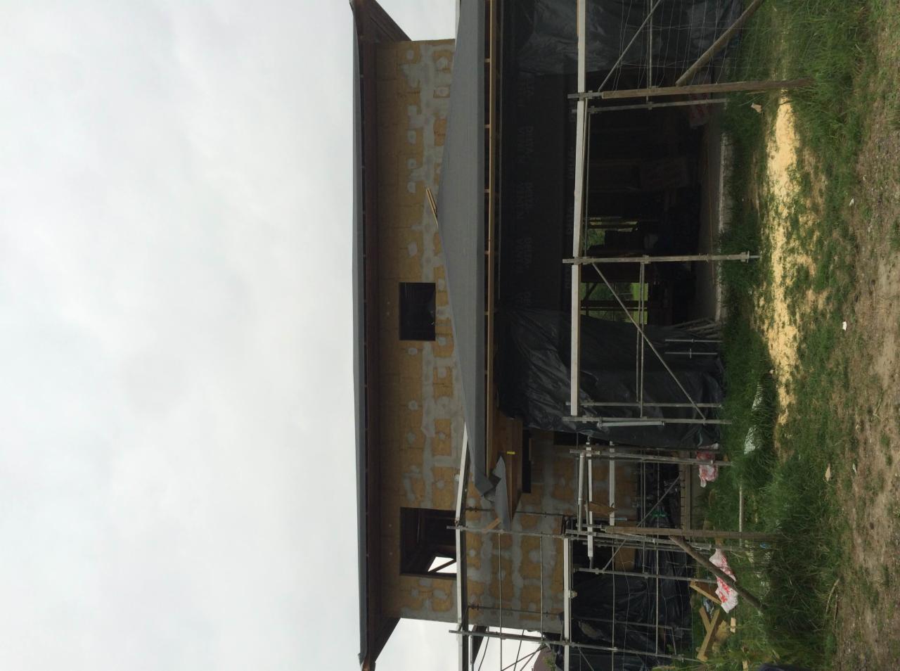 Budowa domu: ściana z widoczną izolacją, częściowo pokryta czarną folią, rusztowania w trakcie montażu, pochmurne niebo.