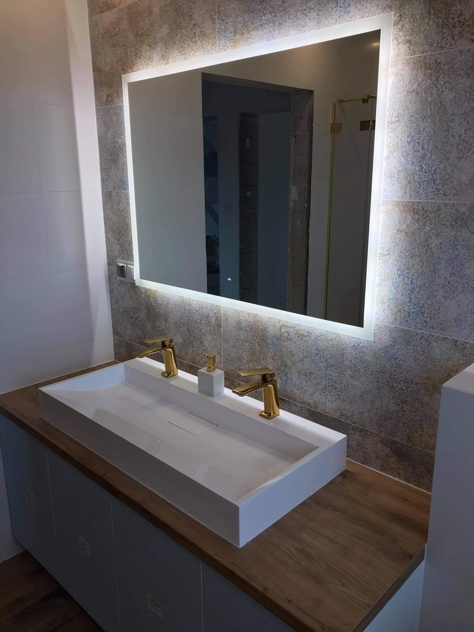 Nowoczesna łazienka z podświetlanym lustrem, białą umywalką nablatową z dwoma złotymi bateriami, blat drewnopodobny i dekoracyjne płytki ścienne z wzorem. W lustrze odbija się częściowo wykończone...