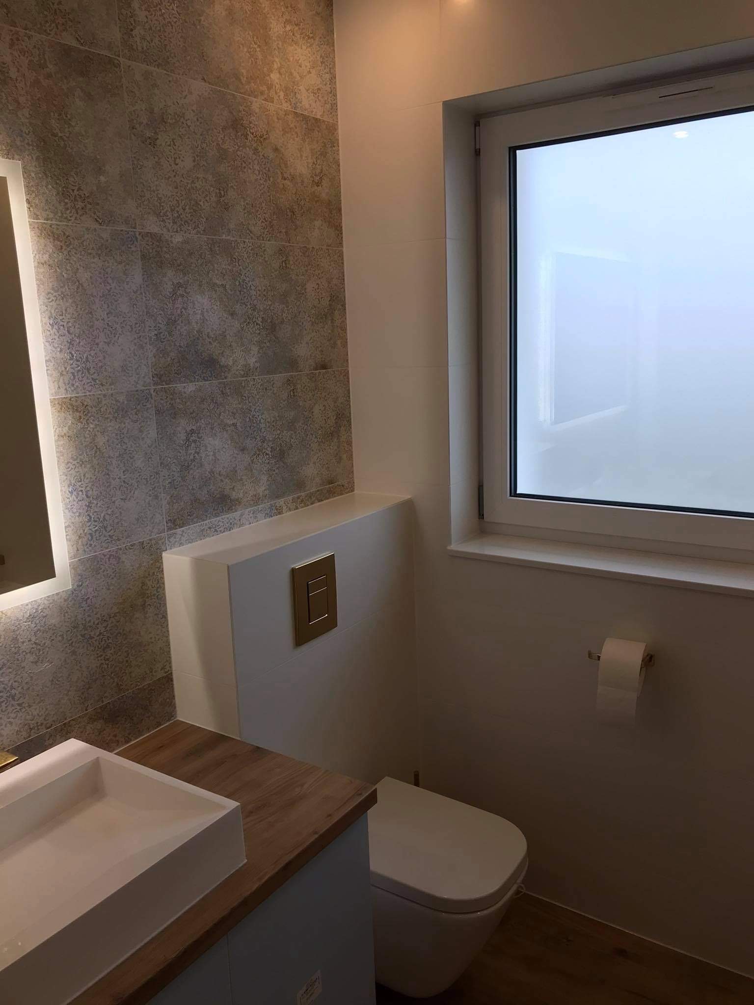 Nowoczesna łazienka z wiszącą miską WC, zabudową stelaża, umywalką nablatową i dekoracyjnymi płytkami ściennymi w odcieniach szarości i beżu.