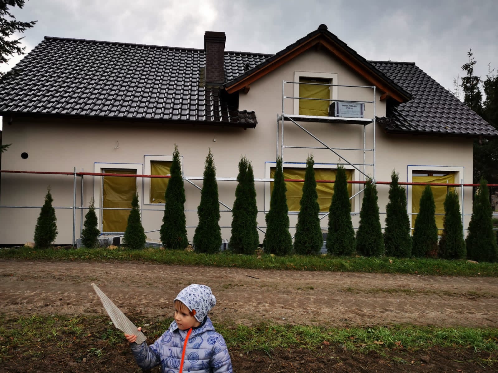 Dom jednorodzinny z nową elewacją w trakcie prac, okna zabezpieczone folią, rusztowanie przy ścianie, rząd tujek przed domem, na pierwszym planie dziecko trzymające fragment materiału izolacyjnego.