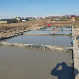 Wylany beton w szalunkach fundamentowych na placu budowy, widoczne wystające rury instalacyjne i sylwetka fotografa w rogu kadru.