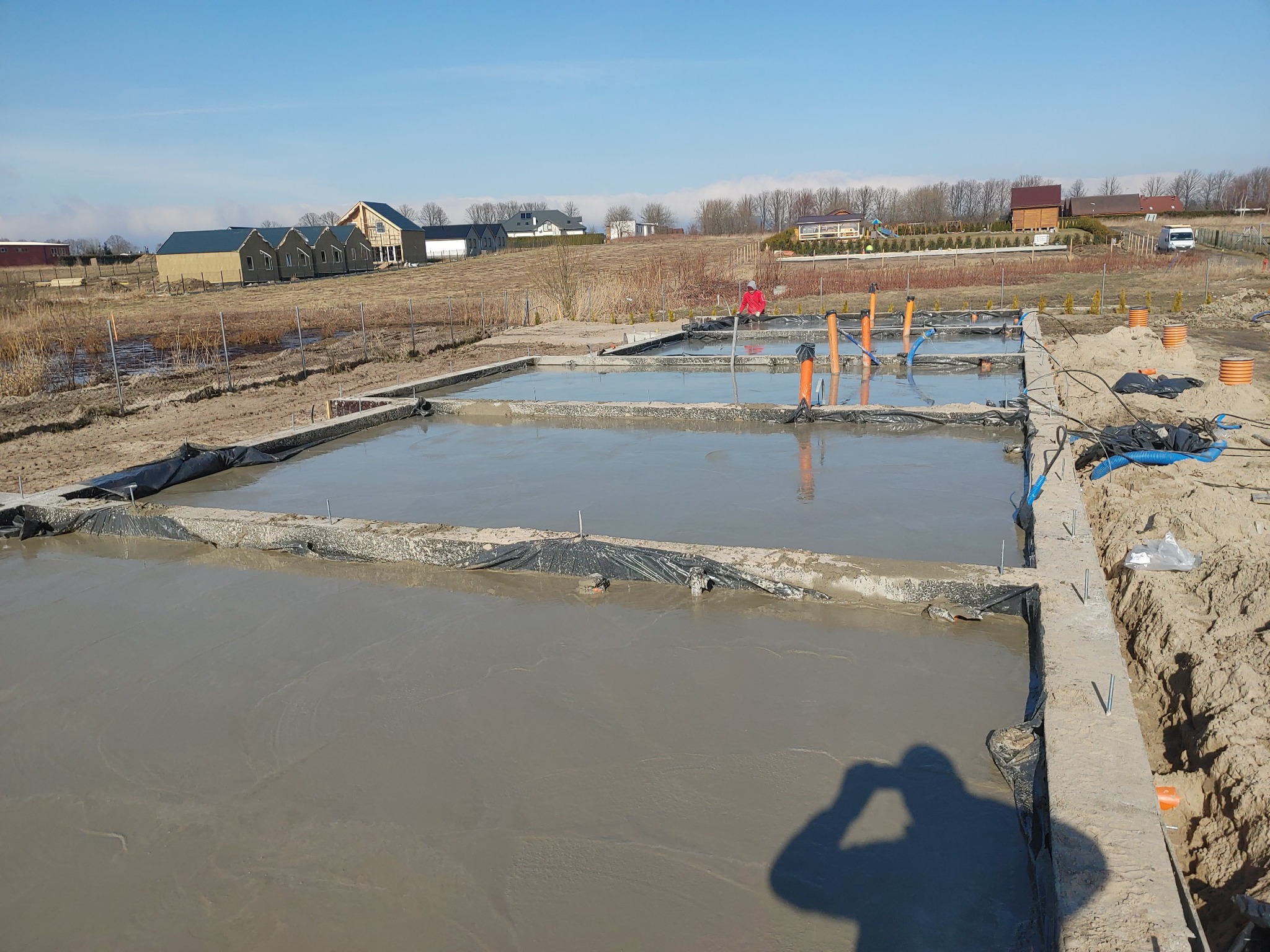 Wylany beton w szalunkach fundamentowych na placu budowy, widoczne wystające rury instalacyjne i sylwetka fotografa w rogu kadru.