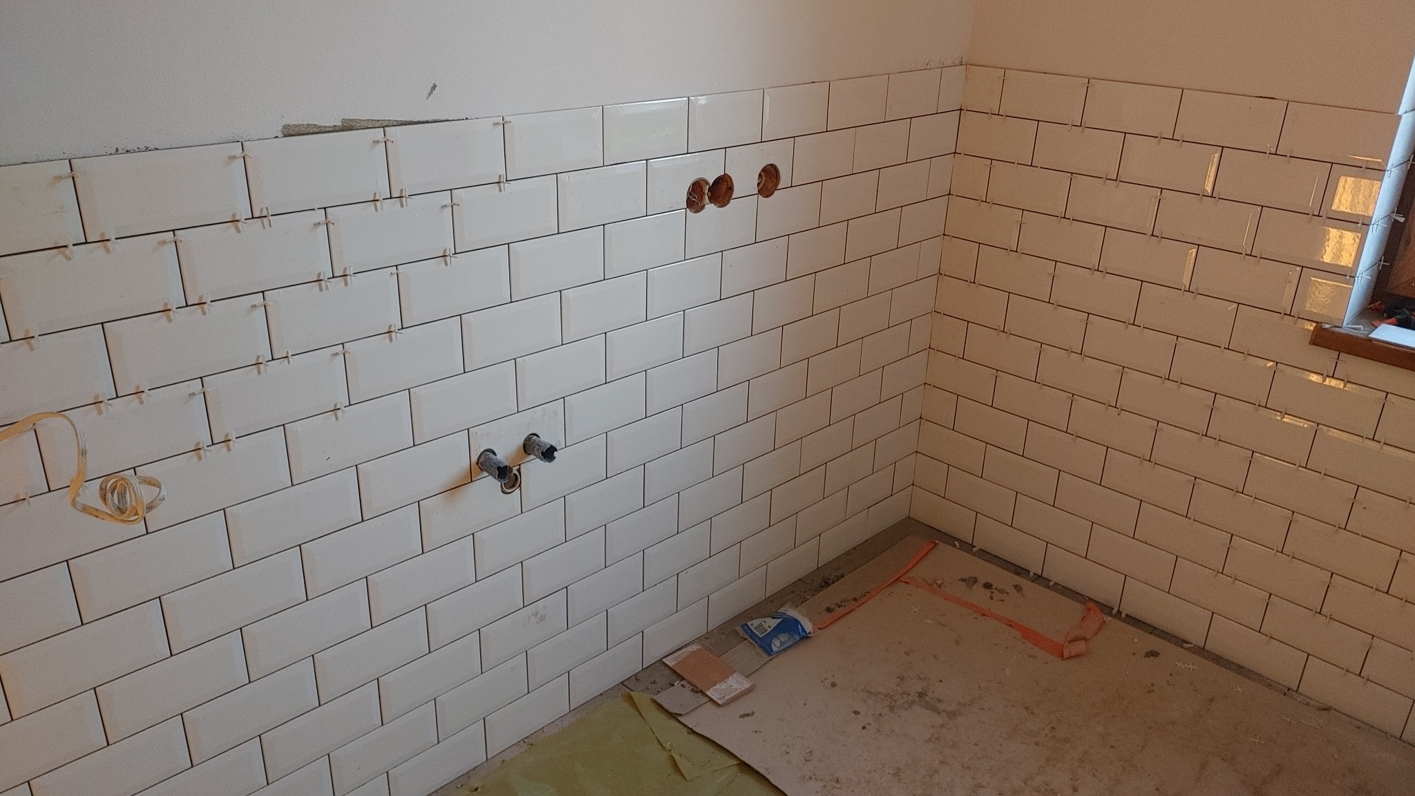 Ściana w trakcie układania białych, prostokątnych płytek, z widocznymi krzyżykami dystansowymi i wystającymi rurami instalacji wodnej, w pomieszczeniu z oknem.