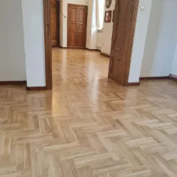 Jasny, lakierowany parkiet ułożony we wzór jodełki francuskiej w przestronnym holu z drewnianymi drzwiami i białymi ścianami.