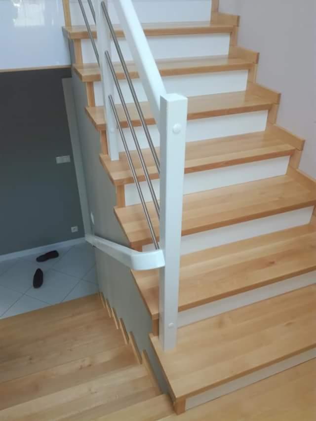 Jasne drewniane schody z białymi podstopnicami i metalową balustradą, widok z dołu na górę, z fragmentem podłogi wyłożonej białymi płytkami i leżącym obok butem.