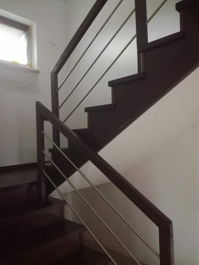 Ciemnobrązowe drewniane schody z metalowymi poręczami w nowoczesnym domu, widoczne fragmenty białych ścian i okna.