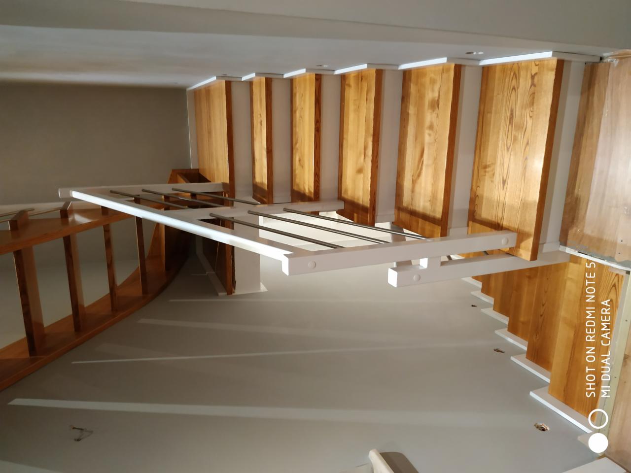 Widok z dołu na drewniane schody z białymi wspornikami i metalowymi poręczami, w trakcie montażu, sufit z widocznymi otworami na oświetlenie.
