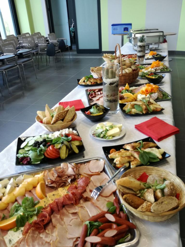 Długi stół z białym obrusem zastawiony różnorodnymi przekąskami, wędlinami, serami, pieczywem i sałatkami, dekorowany kaktusem w szklanym naczyniu, ustawiony w przestronnym pomieszczeniu...