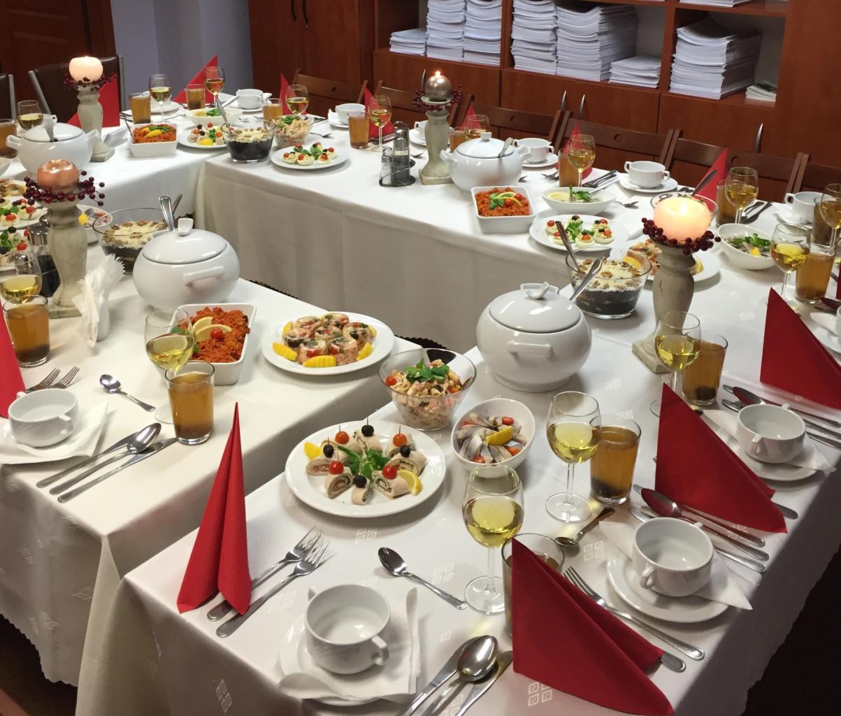 Uroczysty stół z białym obrusem, zastawą i różnorodnymi potrawami: sałatki, rolady, ryby, napoje w szklankach, świece w ozdobnych świecznikach, czerwone serwetki, przygotowany na przyjęcie.