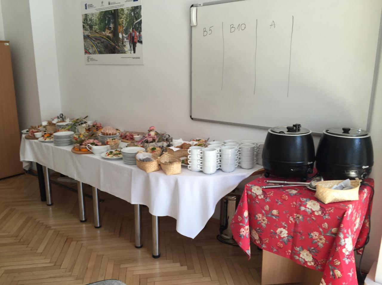 Stół z białym obrusem zastawiony różnorodnymi potrawami, talerzami, koszyczkami z pieczywem i stosem białych filiżanek, obok dwa czarne podgrzewacze do zupy na stole z czerwonym obrusem w kwiaty,...