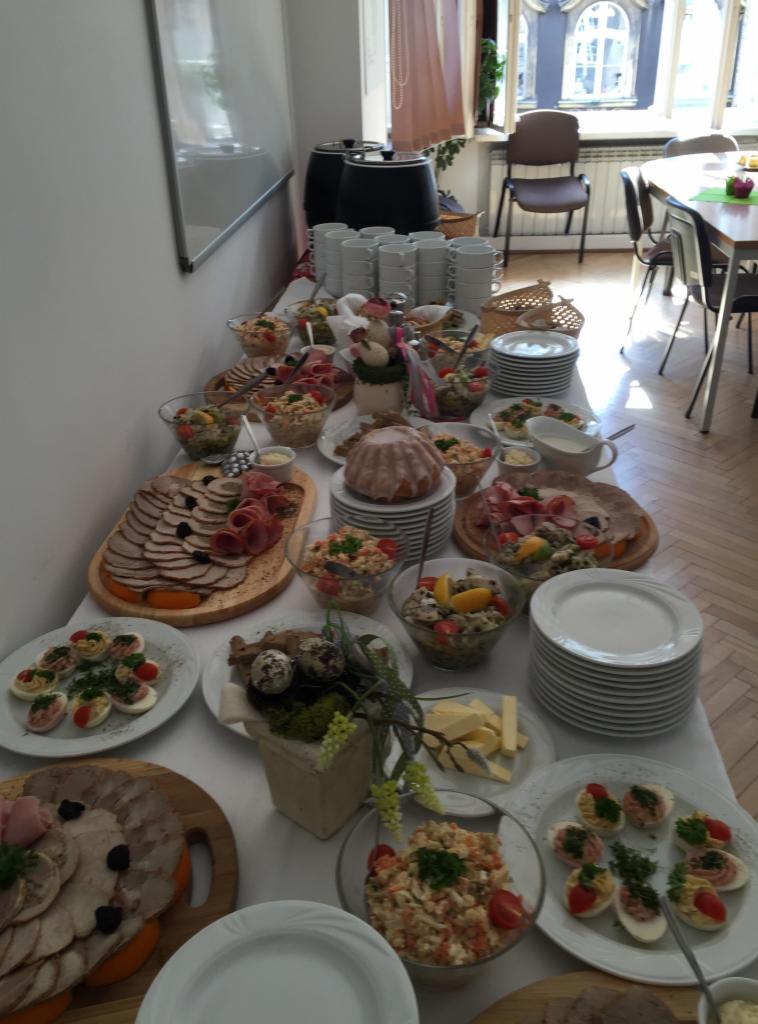 Stół z białym obrusem zastawiony różnorodnymi potrawami: sałatki w szklanych miskach, półmiski z wędlinami, jajka faszerowane, babka wielkanocna, masło w kostkach, stosy białych talerzy, w tle...