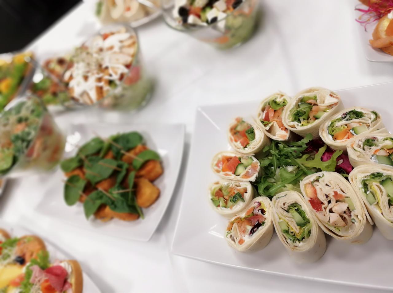 Biały stół z różnorodnymi przekąskami: tortille z łososiem, kurczakiem i warzywami, sałatki w szklanych naczyniach, smażone kąski z zielonymi liśćmi, całość na białym tle.