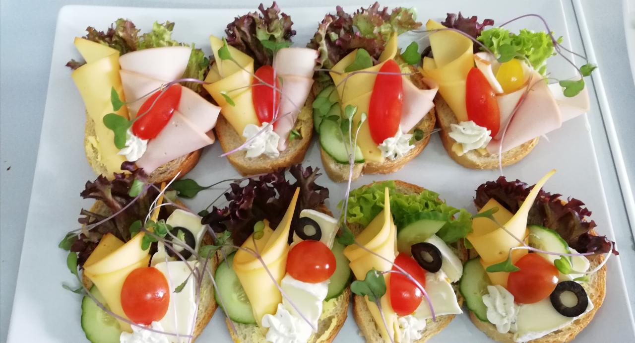Dekoracyjne kanapeczki na białej tacy: pieczywo z kremowym serem, plasterkiem żółtego sera, szynki, ogórka, pomidorkiem cherry, oliwką i listkami sałaty.