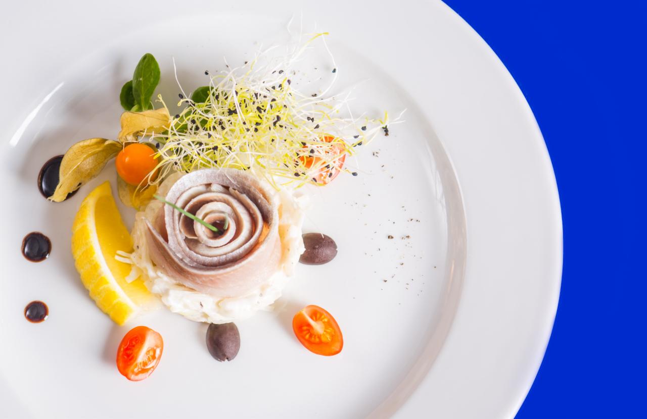 Elegancko podane danie z roladek śledziowych uformowanych w kształt róży na białym talerzu, z dodatkiem cytryny, pomidorków cherry, oliwek, physalis i kiełków, na niebieskim tle.