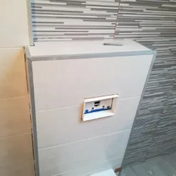 Zabudowa stelaża podtynkowego WC z wykończeniem z płytek ceramicznych w odcieniach bieli i szarości, widoczny fragment muszli klozetowej i przycisk spłukujący.