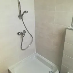 Biały brodzik z zamontowaną baterią i słuchawką prysznicową na tle jasnych, kwadratowych płytek ceramicznych.