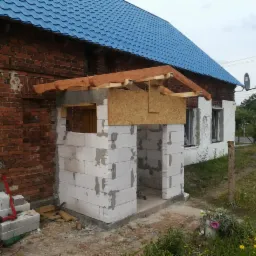 Budowa ganku z bloczków betonowych przed domem z niebieskim dachem, widoczne drewniane elementy konstrukcyjne dachu ganku oraz narzędzia budowlane.