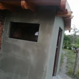 Świeżo otynkowana elewacja budynku gospodarczego z oknem i drzwiami, widoczny fragment ceglanej ściany i drewniane elementy dachu.