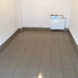 Wnętrze pokoju po remoncie z ułożonymi szarymi płytkami podłogowymi, widoczny grzejnik i wystające przewody elektryczne ze ściany.