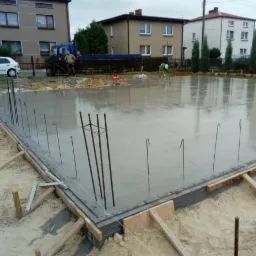 Wylany fundament z wystającymi prętami zbrojeniowymi i drewnianym szalunkiem, widok na budynki mieszkalne i pracowników w tle.