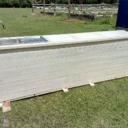 Drewniany bar ogrodowy z metalowym zlewem i kranem, pomalowany na jasny kolor, ustawiony na trawie w plenerze, z przyczepą w tle.