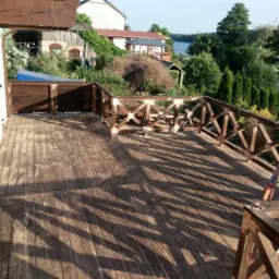 Drewniany taras z balustradą w stylu rustykalnym, widok na jezioro i zieleń, w tle zabudowania gospodarcze.