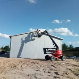 Montaż paneli ściennych na hali magazynowej z użyciem podnośnika koszowego, dwóch pracowników na wysokości, widoczne panele z blachy trapezowej i elementy konstrukcji dachu, teren budowy.