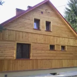 Dom jednorodzinny z drewnianą elewacją w odcieniach miodowego brązu, widok z boku ukazujący liczne okna o ciemnych ramach i czerwony dach.