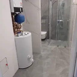 Nowoczesna łazienka z szarymi płytkami imitującymi marmur, kabiną prysznicową ze szklanymi drzwiami, białym bojlerem z niebieskim zbiornikiem wyrównawczym oraz wiszącą toaletą w tle.