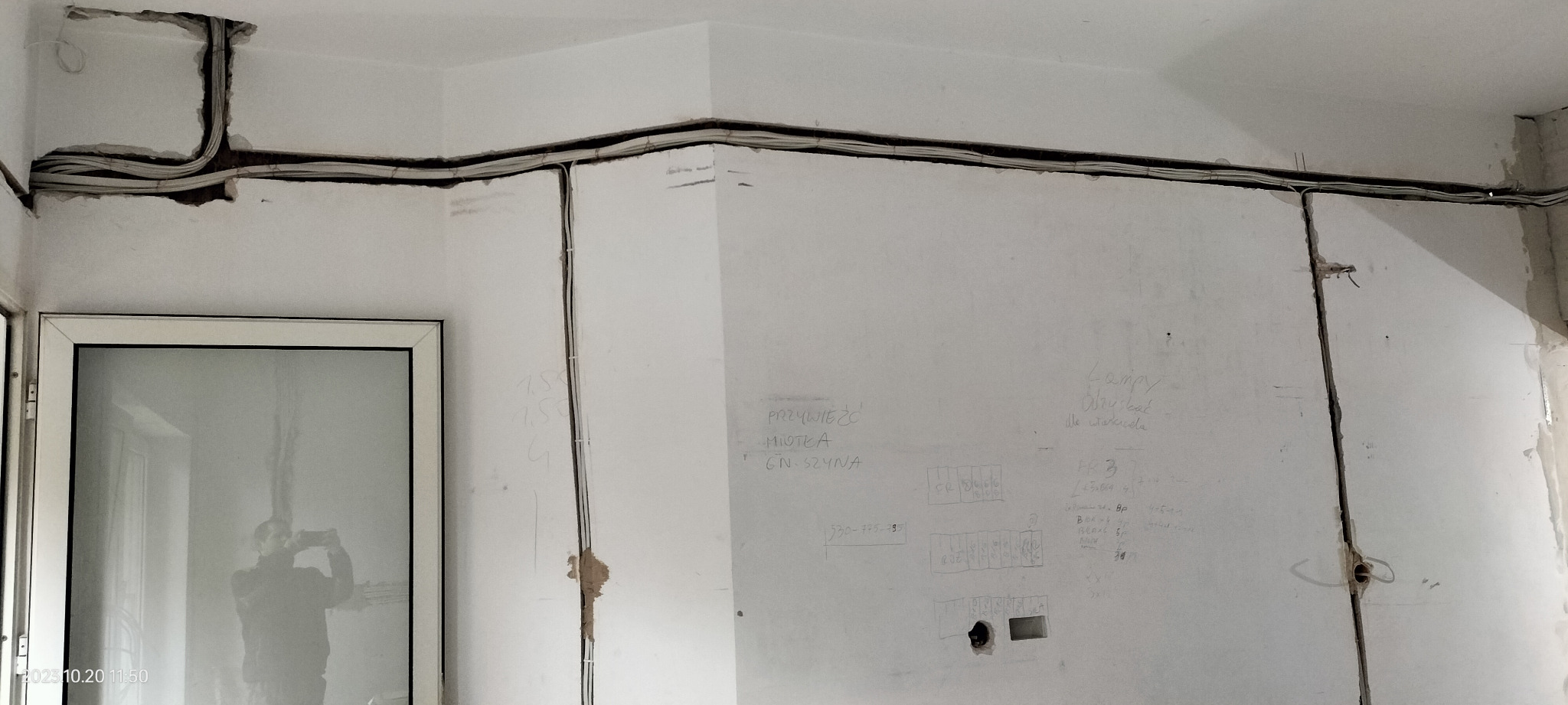 Przewody elektryczne ułożone w bruzdach w ścianie podczas remontu, widoczne notatki i oznaczenia na ścianie, odbicie osoby robiącej zdjęcie w szybie okna.