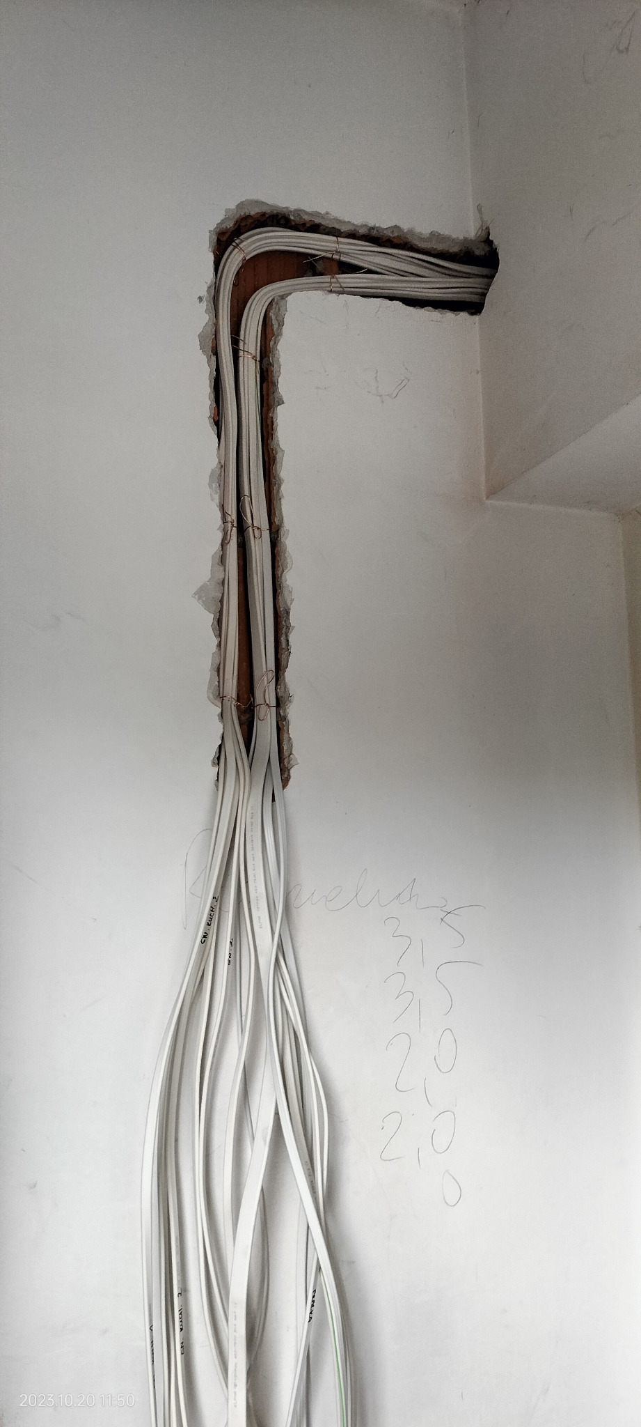 Świeżo położone białe przewody elektryczne wystają ze ściany, zabezpieczone miedzianym drutem, widoczne napisy na przewodach i notatki ołówkiem na ścianie.