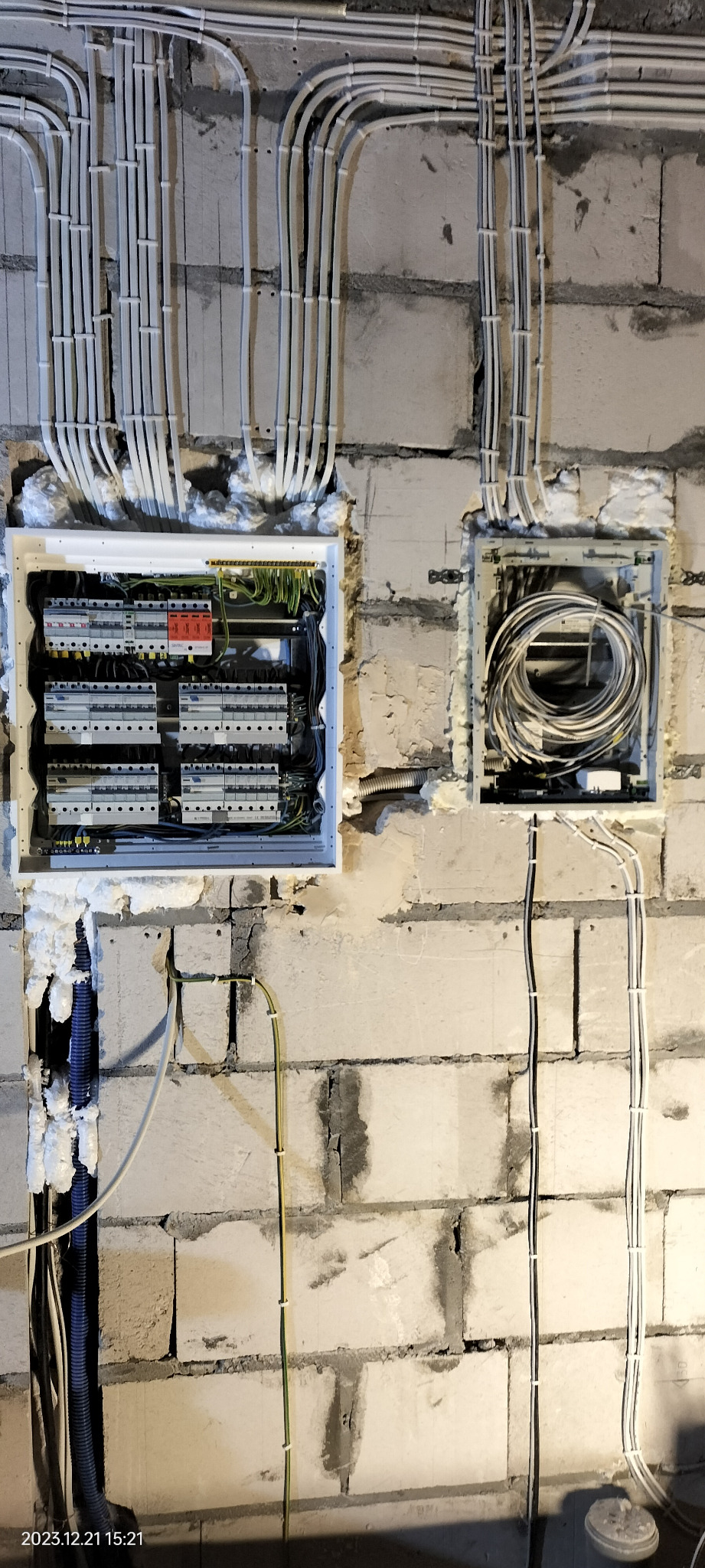 Rozdzielnica elektryczna z zabezpieczeniami i okablowaniem w surowej ścianie z betonu komórkowego, widoczne kable w białych peszlach i puszka z zapasem przewodów.