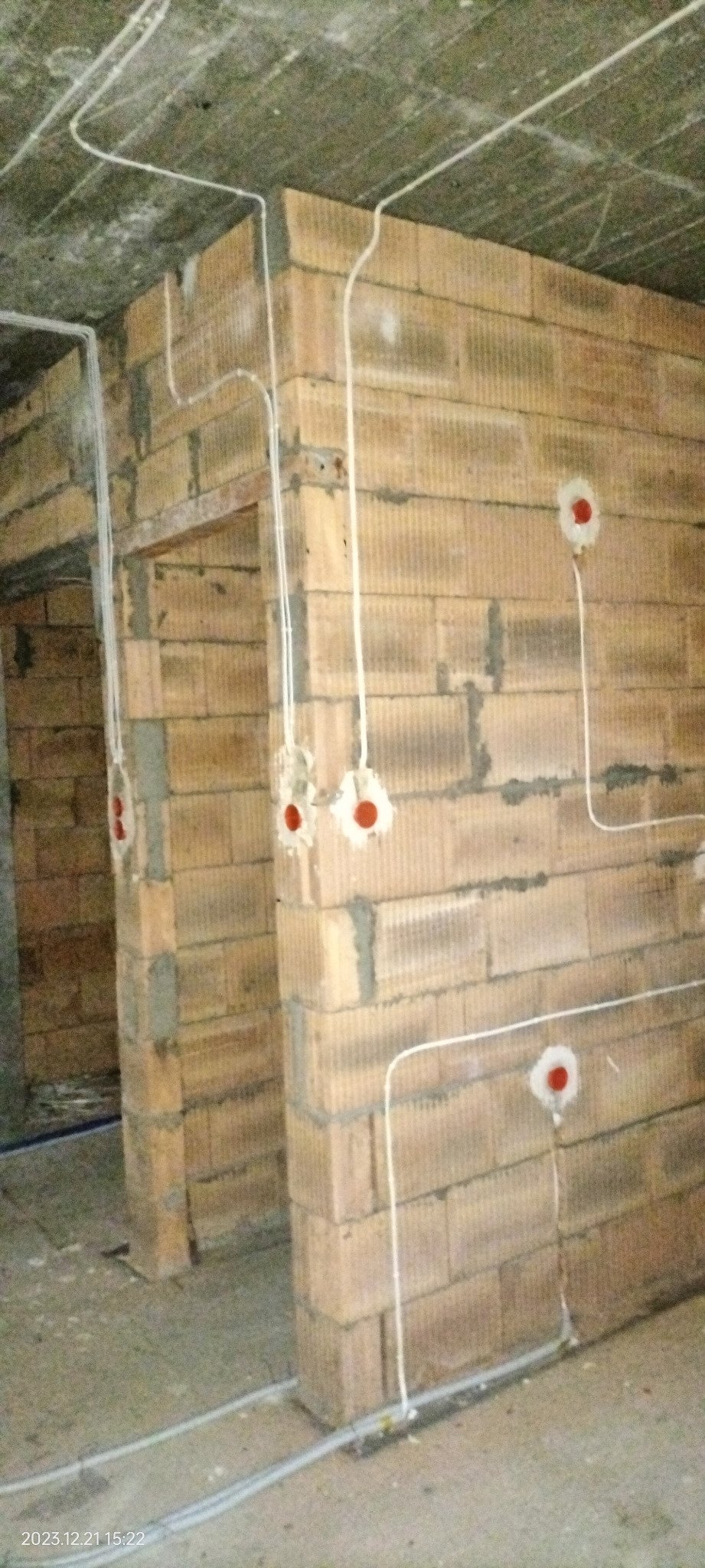 Surowe ściany z cegły ceramicznej z widocznymi przewodami elektrycznymi w białej izolacji, wychodzącymi z czerwonych puszek instalacyjnych; instalacja podtynkowa w trakcie wykonywania.