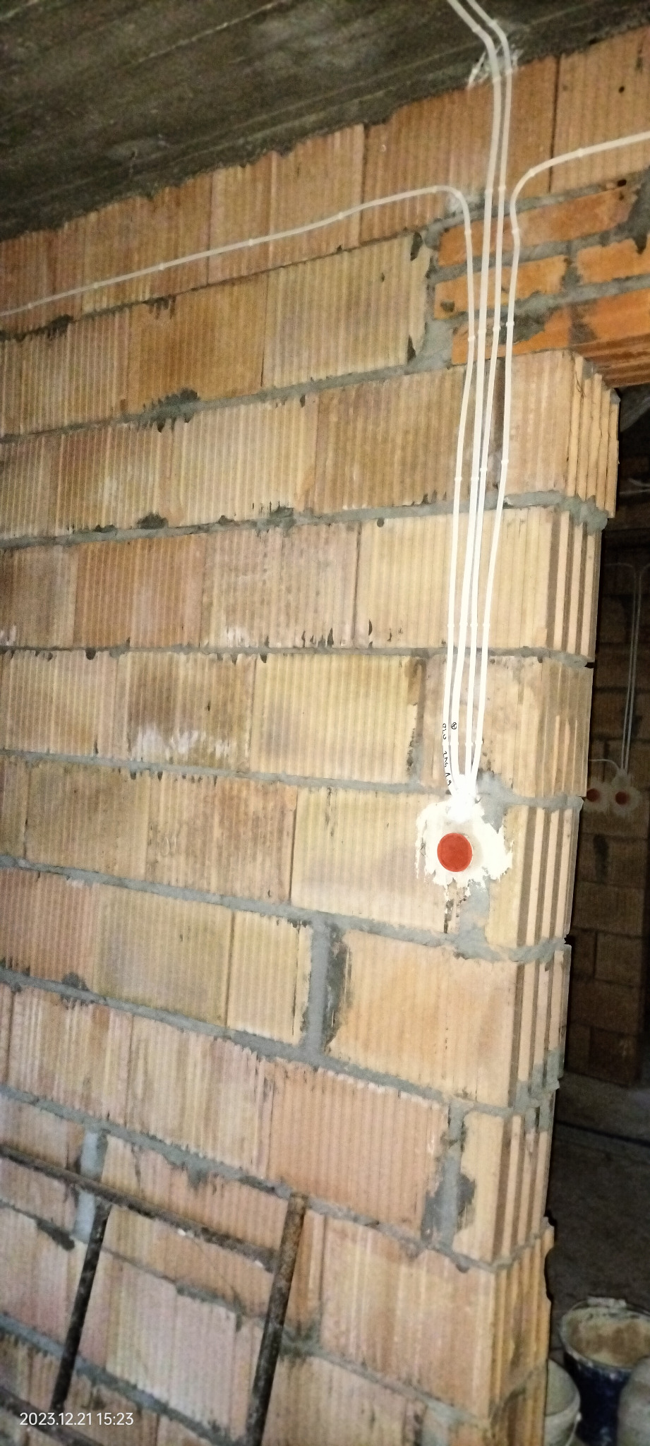 Ściana z cegły dziurawki w trakcie prac instalacyjnych, widoczne kable elektryczne w białej osłonie prowadzone po ścianie i wychodzące z puszki instalacyjnej z czerwonym deklem.