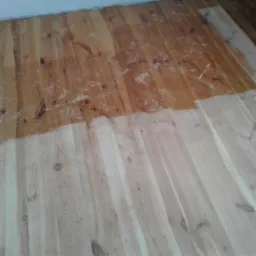 Częściowo wycyklinowana drewniana podłoga z widocznymi śladami pyłu po cyklinowaniu i różnicą w kolorze między wycyklinowaną a niewycyklinowaną częścią.