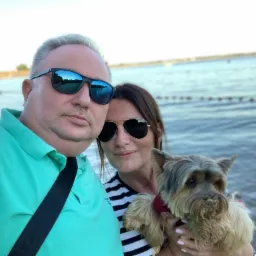 Selfie pary z psem rasy Yorkshire Terrier na tle jeziora w słoneczny dzień; pies ma piasek na pysku.