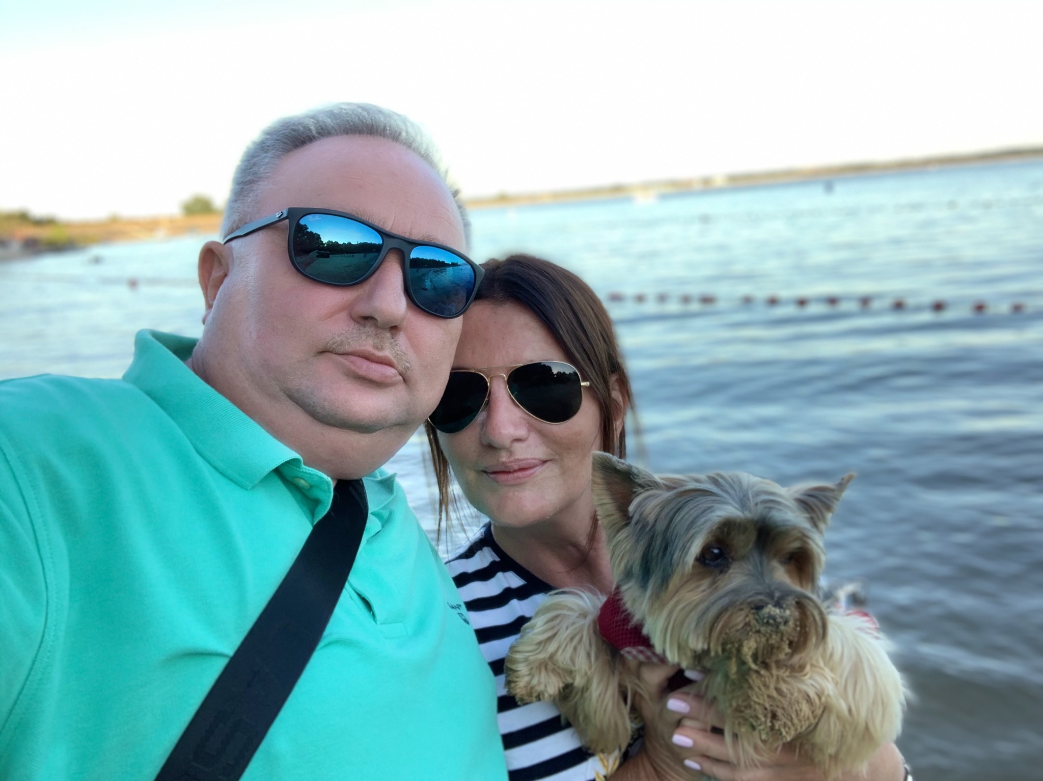 Selfie pary z psem rasy Yorkshire Terrier na tle jeziora w słoneczny dzień; pies ma piasek na pysku.