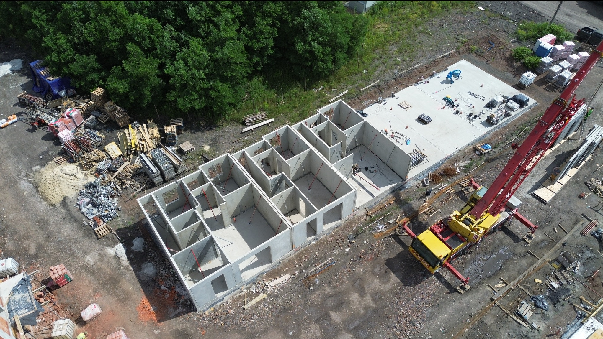 Widok z lotu ptaka na budowę domu z prefabrykatów betonowych. Widać fundamenty, ściany nośne i dźwig. Na placu budowy składowane materiały budowlane i narzędzia.