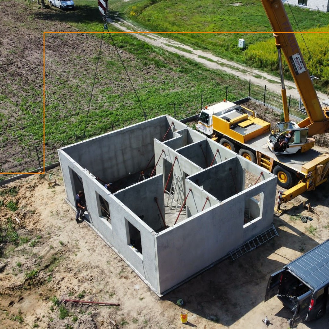 Montaż prefabrykowanego domu z betonu: ściany z otworami okiennymi podnoszone przez dźwig na placu budowy. Widok z góry, częściowo widoczny samochód dostawczy.