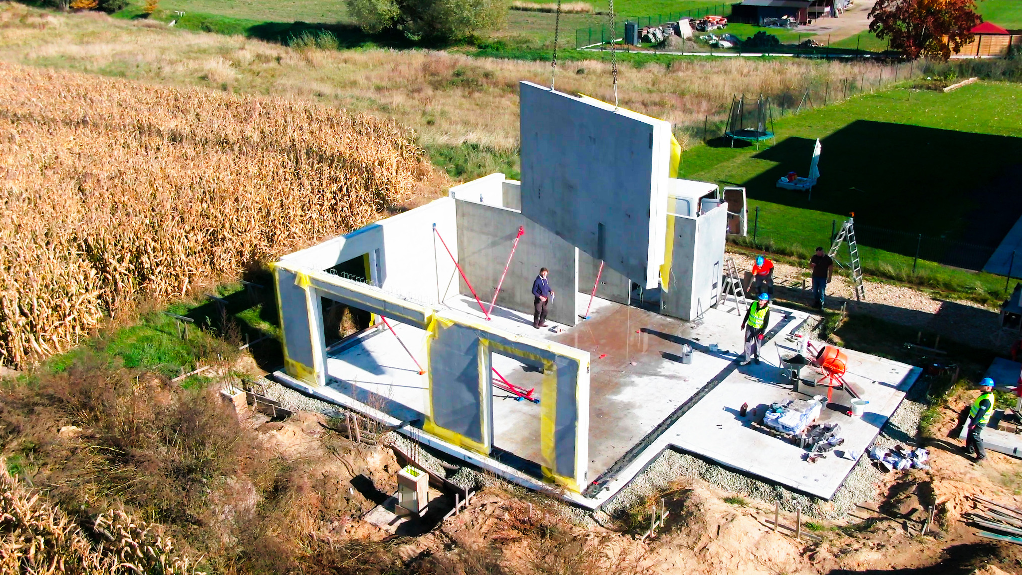 Montaż ścian domu modułowego na fundamencie z użyciem dźwigu. Widoczni pracownicy w kaskach i kamizelkach. W tle pole i zabudowania.