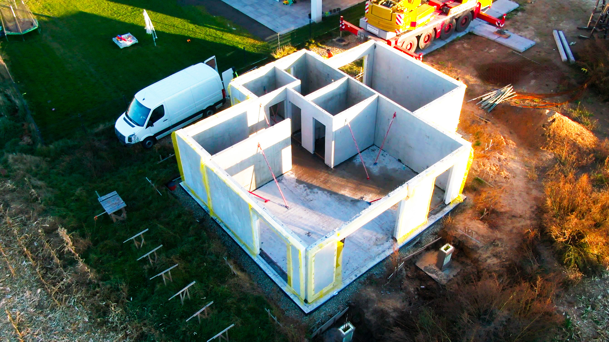 Budowa domu z betonu komórkowego, widok z drona na fundamenty i ściany parteru, z żółtą izolacją. Obok dźwig i biały bus.