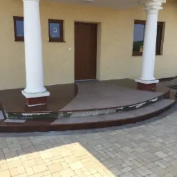 Wejście do domu z kolumnami i schodami wyłożonymi brązowym kamieniem, widoczne niedokończone wykończenie stopni.