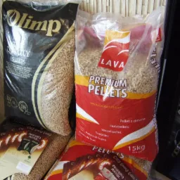 Stos worków z pelletem drzewnym różnych marek, w tym Olimp i Lava, ułożonych w sklepie obok regału z elementami instalacji hydraulicznych.