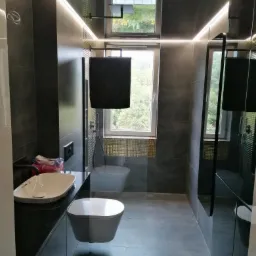 Nowoczesna łazienka z szarymi płytkami, podwieszanym WC i umywalką nablatową, oświetlenie LED i okno z widokiem na zieleń.