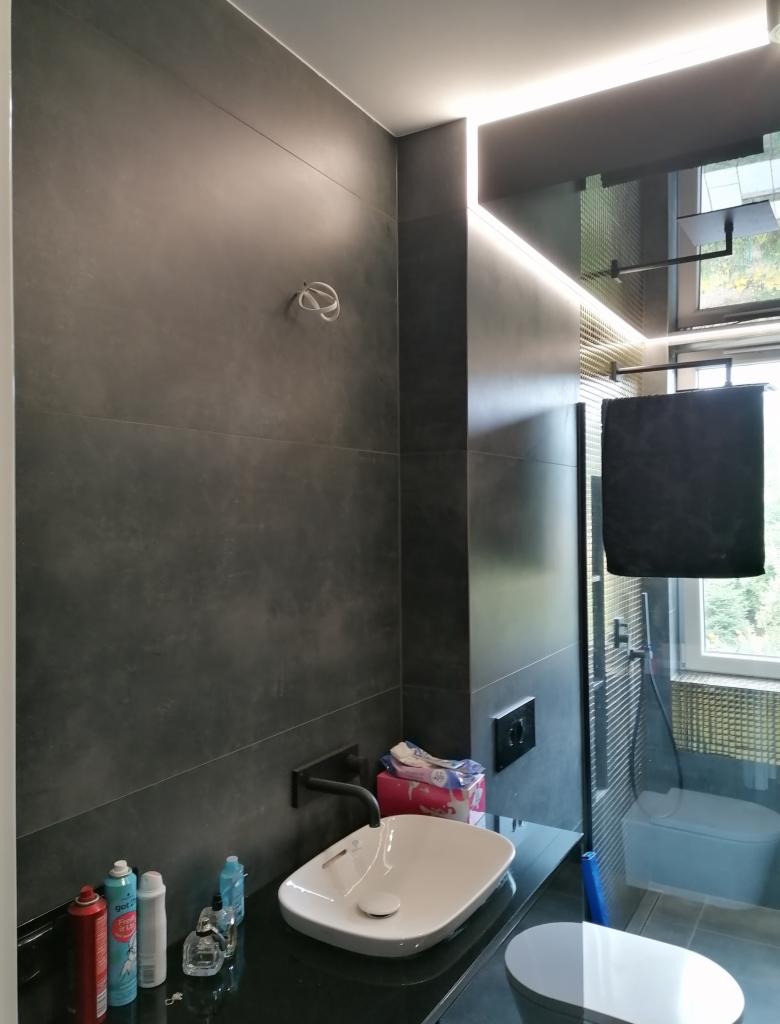 Nowoczesna łazienka z ciemnoszarymi płytkami, podświetleniem LED i białą umywalką nablatową na czarnym blacie. Widoczne kosmetyki i akcesoria łazienkowe. W tle kabina prysznicowa z widokiem...