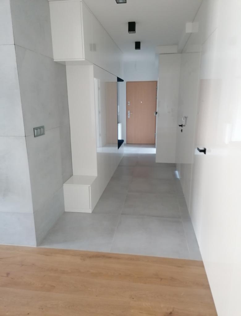 Nowoczesny przedpokój z szarymi płytkami podłogowymi, białymi ścianami z połyskiem, zabudową meblową z szafkami i siedziskiem oraz drewnianą podłogą w strefie wejścia.