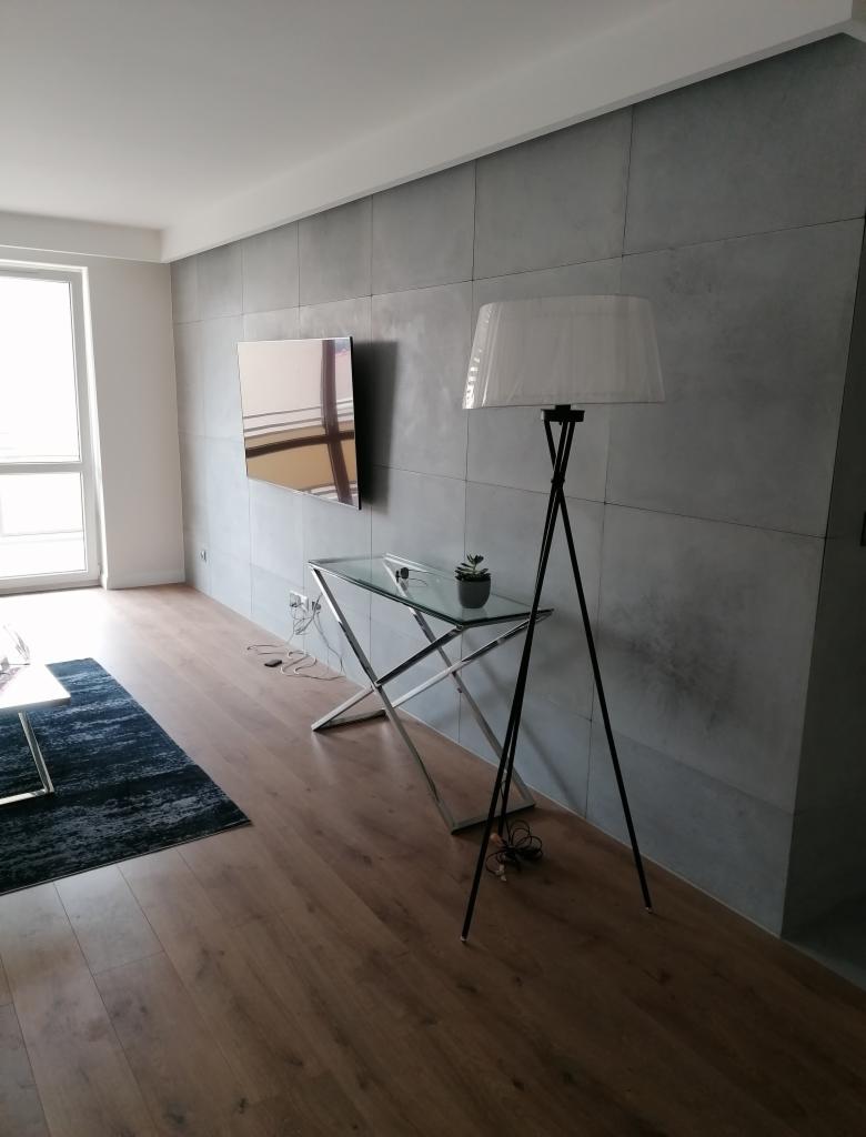 Nowoczesne wnętrze z szarą ścianą wykończoną płytami imitującymi beton, telewizorem na ścianie, szklanym stolikiem z metalowymi nogami, wysoką lampą z abażurem i ciemnym dywanem.